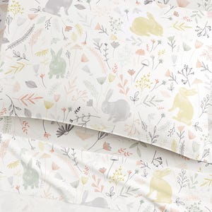 Op de afbeelding: Witte beddengoedset met een herhalend patroon van konijntjes, bloemen en bladeren in zachte pastelkleuren. Het ontwerp bevat grijze, gele en groene konijntjes, met roze, perzik en grijze bloemelementen. Het algemene thema is lente of Pasen.