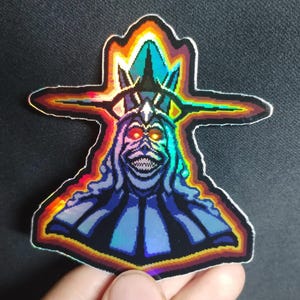 May include: Holographic sticker featuring a pixelated illustration of a spectral figure with a crown and glowing red eyes. The design has a rainbow gradient and a sinister grin, with a black outline.