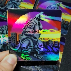 May include: Four holographic stickers featuring a pixelated monster with a beam of light shooting from its mouth. The monster is standing in front of a city skyline. The stickers are iridescent and have a rainbow effect.