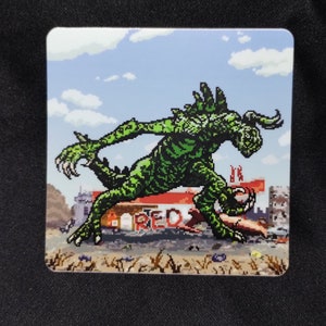 May include: A green pixelated monster with sharp claws and horns is attacking a red building with the word "RED" on it. The monster is in a destroyed city setting with a blue sky and white clouds in the background.