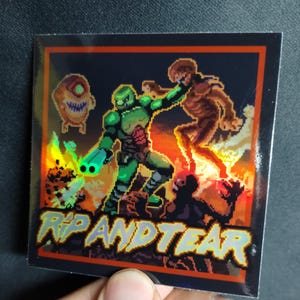 Puede incluir: Adhesivo cuadrado con arte pixelado de una figura verde blindada luchando contra demonios entre llamas. El texto "RIP AND TEAR" está en amarillo y naranja. El adhesivo tiene un borde negro y un efecto holográfico.