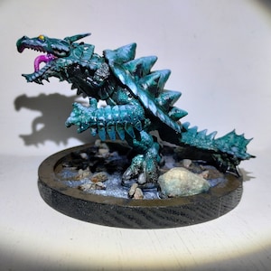 May include: A teal green and black painted miniature dragon figurine with a spiky shell and a pink tongue. The dragon is standing on a round wooden base with a brown and gray textured surface.