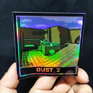 May include: Holographic sticker showing a pixelated video game level with the text "DUST_2" in orange. The sticker has a black border and is held by a hand against a dark background. The image depicts a desert-like environment.