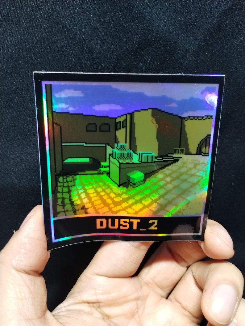 May include: Holographic sticker showing a pixelated video game level with the text "DUST_2" in orange. The sticker has a black border and is held by a hand against a dark background. The image depicts a desert-like environment.