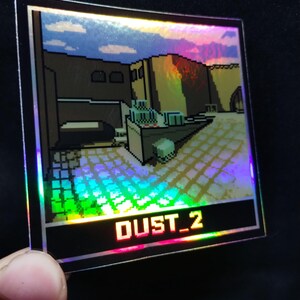 May include: Square holographic sticker with a pixelated desert landscape design. The sticker has a black border and the text "DUST_2" in red. The image has a rainbow effect, with vibrant colours.
