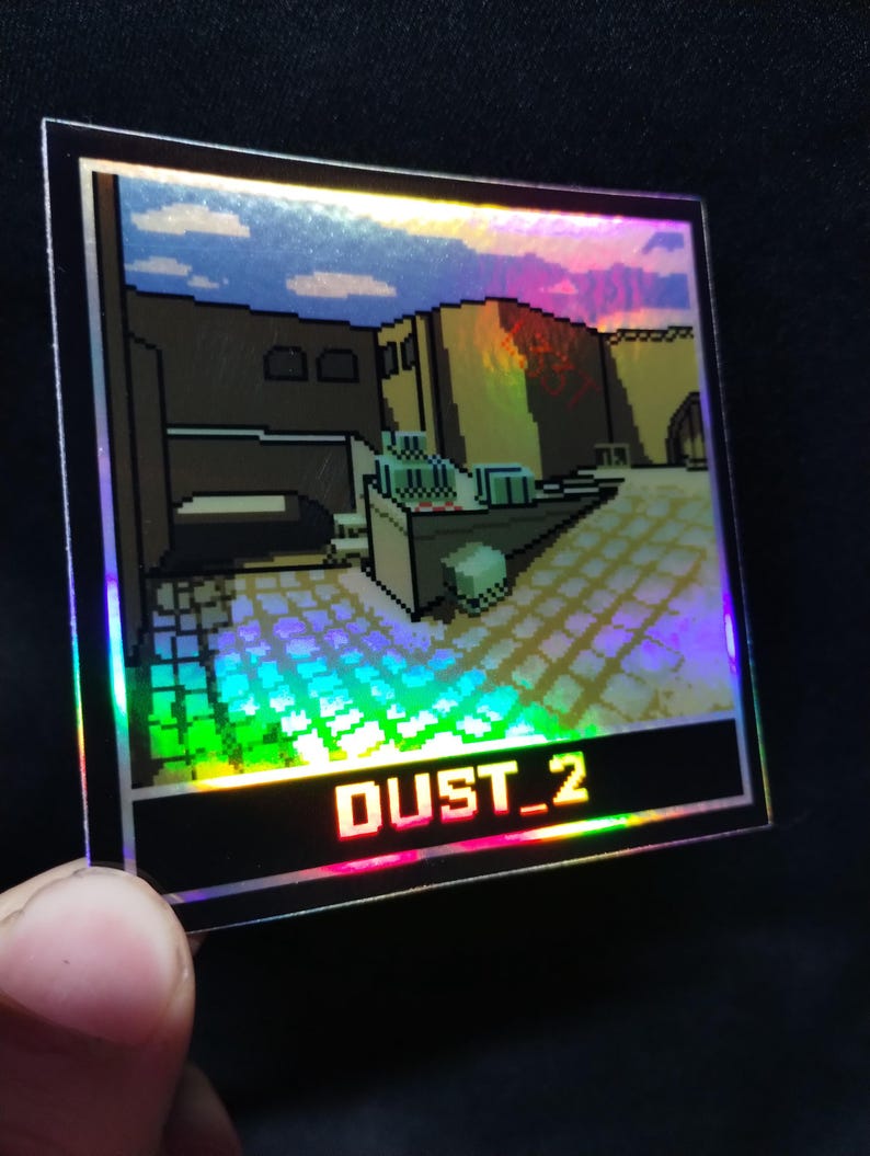May include: Square holographic sticker with a pixelated desert landscape design. The sticker has a black border and the text "DUST_2" in red. The image has a rainbow effect, with vibrant colours.