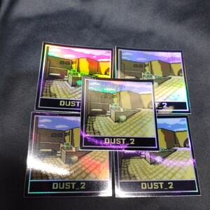 May include: Five holographic stickers displaying a pixelated desert scene with the text "DUST 2". The stickers have a reflective, iridescent finish, with a black border. The background is a dark, textured surface.