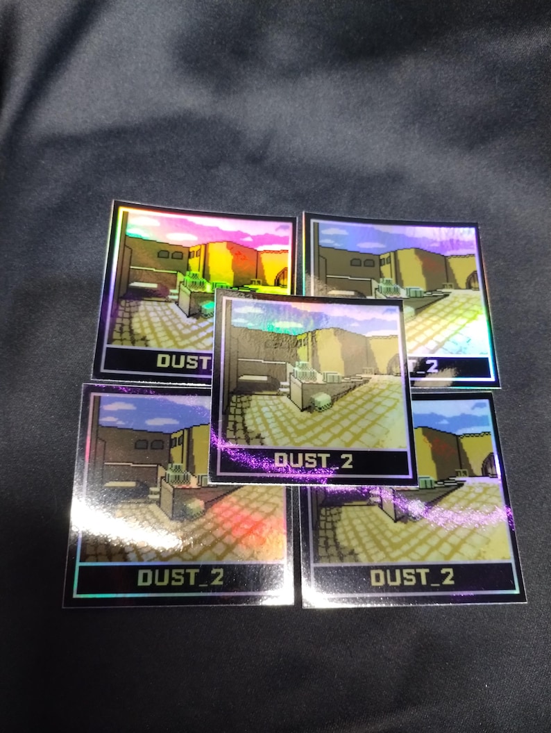 May include: Five holographic stickers displaying a pixelated desert scene with the text "DUST 2". The stickers have a reflective, iridescent finish, with a black border. The background is a dark, textured surface.