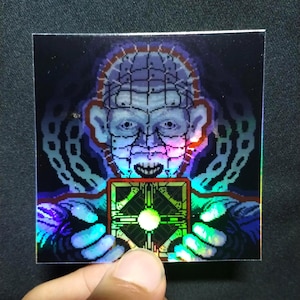 May include: Square holographic sticker featuring a pixelated image of a character holding a puzzle box. The character has a pale face with a grid-like pattern and pins. The puzzle box is green and gold with a central light. The sticker has a dark background with chains.