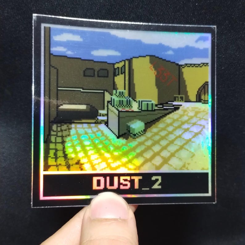 May include: Holographic sticker with a pixelated desert scene, buildings, and a blue sky. The sticker reads "DUST_2" in white text at the bottom. It has a reflective, rainbow effect.