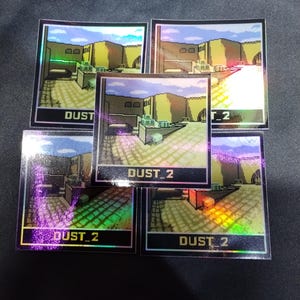 May include: Five holographic stickers displaying a pixelated landscape with the text "DUST_2". The stickers have an iridescent finish, with a colour palette of yellow, brown, and blue.
