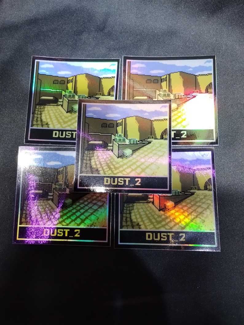 May include: Five holographic stickers displaying a pixelated landscape with the text "DUST_2". The stickers have an iridescent finish, with a colour palette of yellow, brown, and blue.