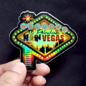 May include: A holographic sticker featuring a neon sign design. The sign reads "Welcome to Fabulous Neon Vegas" in a retro font. The sticker has a black border and a colorful, shimmering effect.