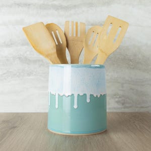 May include: A cylindrical ceramic utensil holder with a teal base and a white drip glaze pattern at the top. The holder contains five light-colored bamboo kitchen utensils, including spatulas, a fork, and slotted spoons, arranged upright.