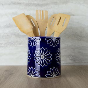 May include: A cylindrical ceramic utensil holder in deep blue with a repeating white starburst or floral pattern. The container is filled with five light-colored bamboo kitchen utensils, including spatulas and a fork, arranged in an upright position.