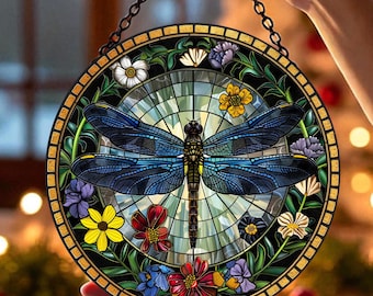 May include: Round stained glass window art featuring a blue dragonfly with intricate wing details. The dragonfly is surrounded by colorful flowers and greenery. A black chain is attached for hanging. The artwork is held up by a person's hands.
