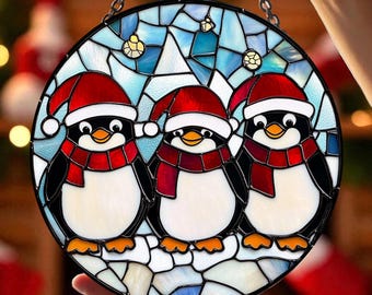 May include: A round stained glass decoration featuring three penguins wearing red Santa hats and scarves. The penguins are set against a blue and white background with a chain for hanging. A festive holiday decoration.