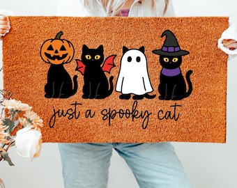 May include: A rectangular doormat with a Halloween theme. It features four cartoon figures: a jack-o'-lantern, a black cat with bat wings, a ghost, and a black cat wearing a witch hat. The text "just a spooky cat" is written below the figures.