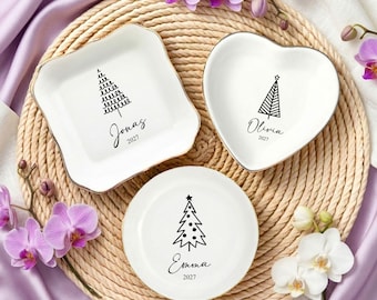 May include: Three white ceramic trinket dishes with gold-toned rims. One square dish features a tree graphic and the name Jonas 2027. One heart-shaped dish features a tree graphic and the name Olivia 2027. One round dish features a tree graphic and the name Emma 2027.