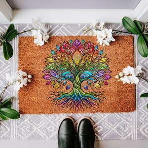 May include: A rectangular doormat with a vibrant, colorful tree of life design. The tree features a rainbow of colors, including red, orange, yellow, green, blue, and purple. The mat is made of a natural brown material and is placed on a gray patterned surface.