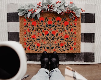 May include: A brown coir doormat with a red floral design and dark green leaves. The mat is placed on a patterned rug with a black and white buffalo check pattern. A sprig of red berries and green leaves sits on top.