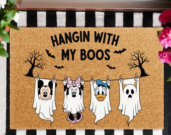 May include: A rectangular doormat with the text "HANGIN WITH MY BOOS" and illustrations of Mickey Mouse, Minnie Mouse, Donald Duck, and a ghost, all dressed as ghosts. The mat has a brown background with a black and white striped border.