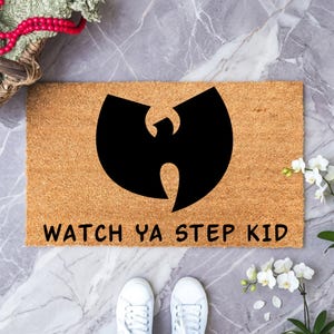 May include: A rectangular tan coir doormat featuring a large black Wu-Tang Clan logo centered in the middle. Below the logo, the text "WATCH YA STEP KID" is printed in bold black capital letters. The mat has a coarse, natural fiber texture.