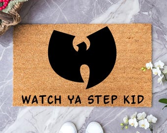 May include: A rectangular tan coir doormat featuring a large black Wu-Tang Clan logo centered in the middle. Below the logo, the text "WATCH YA STEP KID" is printed in bold black capital letters. The mat has a coarse, natural fiber texture.
