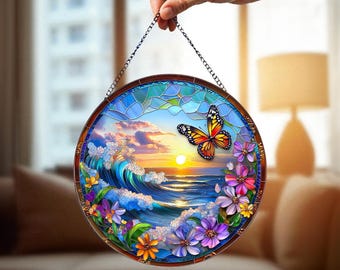 May include: Round stained glass window art featuring a sunset over the ocean with a butterfly and colorful flowers. The artwork is framed with a dark brown border and suspended by a chain.