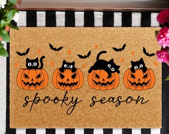 May include: A rectangular doormat with a Halloween theme. It features four orange jack-o'-lanterns with black cats on top, black bats, and the words "spooky season" in a script font. The mat has a natural fiber appearance.