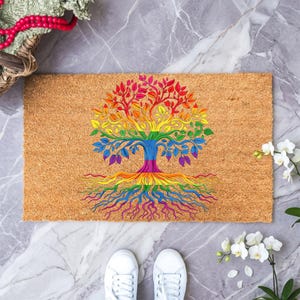May include: A rectangular doormat made of natural coir fiber. It features a vibrant rainbow-colored tree design, with roots, trunk, branches, and leaves in a spectrum of colors. The mat is placed on a marble surface, with white sneakers visible.