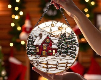May include: Round stained glass Christmas ornament featuring a red house with snow-covered roof and surrounding trees. The ornament has a white and clear background with a metal chain for hanging. The house has lit windows and a white picket fence.