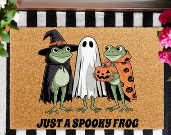 May include: A Halloween-themed doormat featuring three cartoon frogs dressed in costumes. One frog wears a witch hat and cape, another is a ghost, and the third holds a jack-o'-lantern. The text "JUST A SPOOKY FROG" is at the bottom.