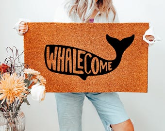 May include: A rectangular doormat with a brown coir surface. A black whale silhouette with the word "WHALECOME" is centered on the mat. The mat is held up by a person. A vase of flowers is in the background.