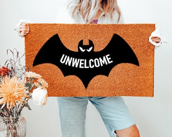 May include: A rectangular doormat with a brown coir surface. A black bat silhouette with the word "UNWELCOME" in white is centered on the mat. The mat is held up by a person. A vase of flowers is in the background.