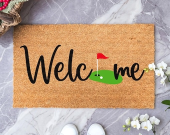 May include: A rectangular tan coir doormat featuring the black cursive text "Welcome" with the letter "c" replaced by a green golf putting green graphic, a white golf ball, and a red flag on a pole, printed in the center of the mat.