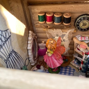 May include: A miniature fairy figurine with pink wings and a pink dress stands in a dollhouse-like setting. The dollhouse is decorated with a sewing theme, including spools of thread, a vintage clock, and a dress form.