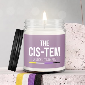 May include: A clear glass jar candle with a lit wick and a black lid. The label is light purple with white text that reads THE CIS-TEM OH LOOK... IT'S ON FIRE, featuring a horizontal stripe in yellow, white, purple, and black at the bottom.