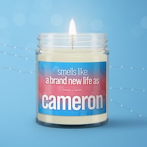 May include: A white candle in a glass jar with a blue and pink label that says "smells like a brand new life as cameron".