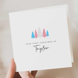 May include: A white Christmas card with the text "YOUR FIRST CHRISTMAS AS Taylor". The card features a design of stylized Christmas trees in blue, pink, and gray. The card is folded and held by a hand.