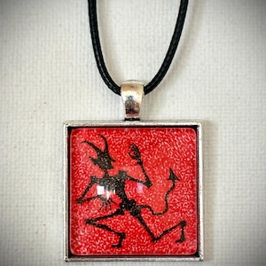 Peut inclure: Un pendentif carré monté sur un cordon noir, présentant la silhouette noire d'une créature cornue et dotée d'une queue sur un fond rouge texturé. Le motif est enchâssé dans un cadre en métal argenté, offrant un contraste visuel marqué.