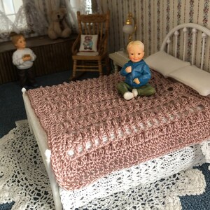 May include: A dollhouse bed with a pink crocheted blanket and a small doll sitting on the bed.