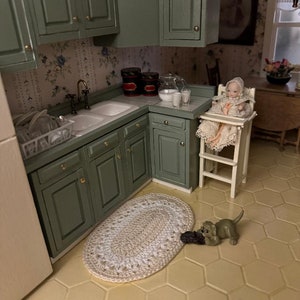 Puede incluir: Una cocina de casa de muñecas con armarios verdes, un fregadero blanco y un suelo de baldosas blancas. Una muñeca está sentada en una trona y una pequeña figura de perro está en el suelo. Hay una alfombra de ganchillo en el suelo.