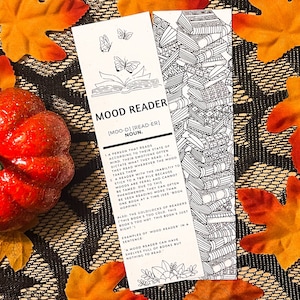 Könnte beinhalten: Ein schwarz-weißes Lesezeichen mit einer Buchillustration und Text, der lautet "MOOD READER [MOO-D] [READ-ER] NOUN. 1. Eine Person, die liest, je nach ihrem Gemütszustand, ihre Emotionen diktieren oft, was sie lesen. 2. Ein Leser, der nicht in der Lage ist, sich an einen Bücherstapel zu halten, weil Stimmungen wild sind und aufgrund dieses Phänomens nicht gezähmt werden können. Sie können oft gesehen werden, wie sie mehr als ein Buch gleichzeitig lesen. Ich sehe ein Buch, das sich moppt. Auch die Goldlöckchen der Leser dieses Buch ist zu kalt dieses Buch ist zu heiß dieses Buch ist genau richtig! Beispiele für einen Stimmungsleser in einem Satz Ein Stimmungsleser kann Regale voller Bücher haben, aber nichts zu lesen."