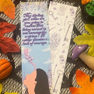 May include: Two decorative bookmarks. One features a woman looking up at a butterfly with the text "My darling girl, when are you going to realize that being normal is not necessarily a virtue? It rather denotes a lack of courage." The other has keys and lavender.