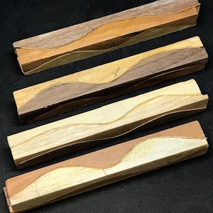 Op de afbeelding: Vier rechthoekige houten stukken met een golvend, ingelegd ontwerp. De houttinten variëren van licht naar donker, inclusief tinten bruin, beige en lichtbruin. De stukken zijn parallel gerangschikt en tonen het vakmanschap en de natuurlijke houtnerf.