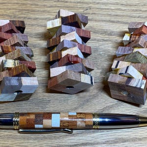 Op de afbeelding: Een houten pen met een mozaïekontwerp, met verschillende houttinten, ligt onder drie stapels houten puzzelstukjes. De puzzelstukjes zijn gemaakt van verschillende houtsoorten en -kleuren, waaronder bruin, beige en rood.