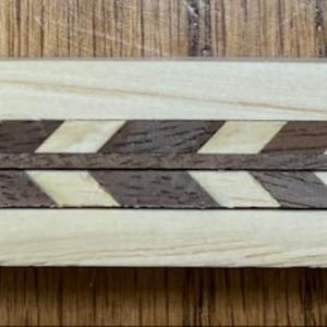 May include: Close-up of a wooden decorative element with a chevron pattern. The design features alternating light and dark wood pieces, creating a contrasting visual effect. The central band is a darker shade, with lighter wood on the top and bottom.