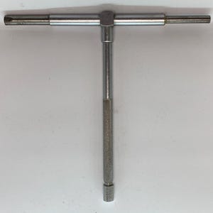 May include: A silver-colored T-handle wrench with a textured grip. The tool has a T-shape with a horizontal bar and a vertical shaft. The handle is knurled for a secure grip. The wrench is designed for tightening or loosening screws or bolts.