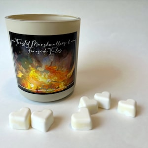 May include: A beige candle holder with a black label featuring the text "Toasted Marshmallows & Fireside Tales" and a campfire scene. Six heart-shaped wax melts are scattered around the candle.